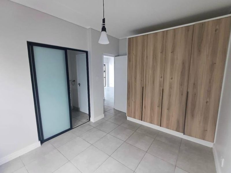 A modern, empty bedroom featuring neutral tiled flooring, ample built-in wooden cupboards, and a contemporary frosted glass sliding door.