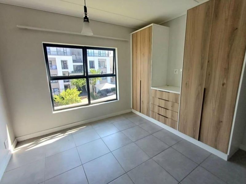 A bright, modern bedroom featuring neutral tiled flooring, large aluminium-framed windows, and ample built-in wooden cabinetry.