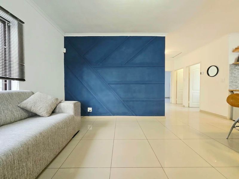Open plan living area featuring large format floor tiles and a striking blue geometric feature wall, leading into a hallway and kitchen space.