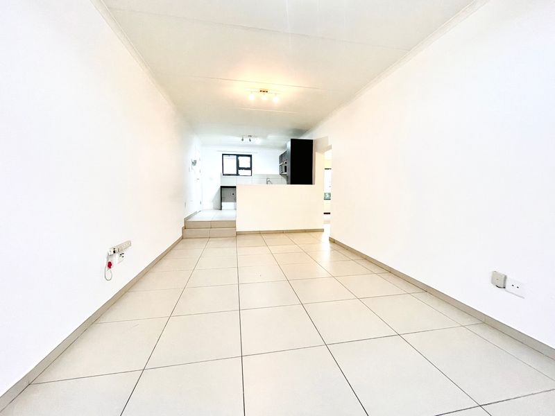 An empty open-plan living and kitchen area featuring neutral tiled flooring, clean white walls, and functional kitchen cabinetry.
