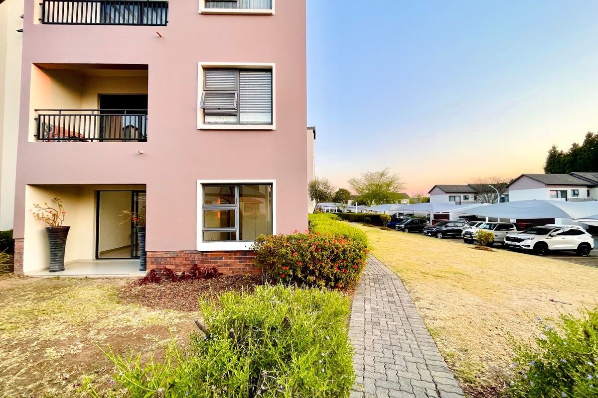 Exterior view of an apartment building featuring a pink facade, private balconies, and a paved walkway leading through the landscaped grounds.