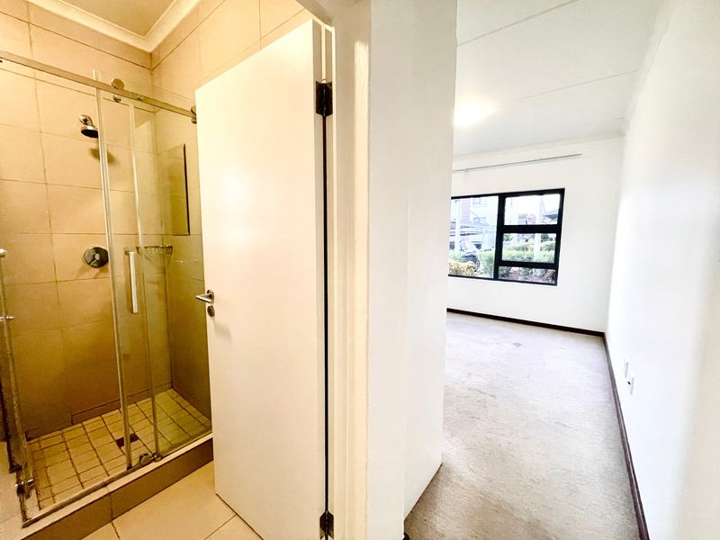 A clean, empty bathroom featuring a glass-enclosed shower with tiled walls, adjacent to a bright, carpeted room with large windows.