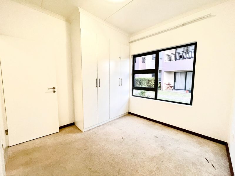 A vacant bedroom featuring built-in cupboards, carpet flooring, and an aluminium-framed window overlooking the complex grounds.