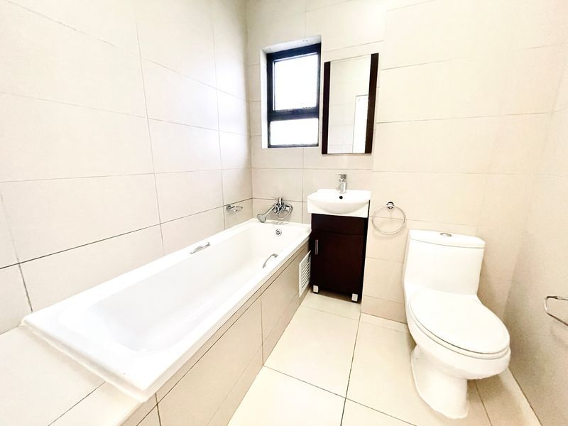 A clean, modern bathroom featuring a built-in bathtub, vanity cabinet with basin, and a standard toilet, finished with neutral floor-to-ceiling wall tiles.