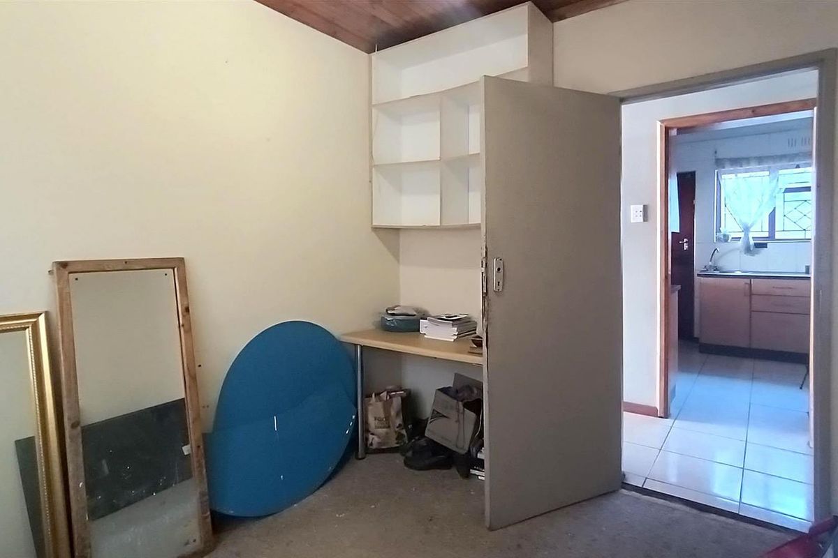 A bedroom featuring built-in shelving and a desk, with a view through to the kitchen area.