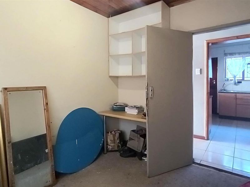 A bedroom featuring built-in shelving and a desk, with a view through to the kitchen area.