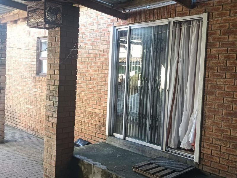 A covered patio area featuring exposed brick walls, a sliding glass door with a security gate, and paved flooring.