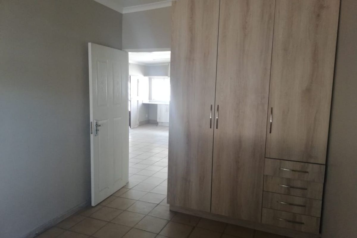 A clean, empty bedroom featuring light wood-finish built-in cupboards, neutral wall tones, and tiled flooring, with a view into an adjacent room.