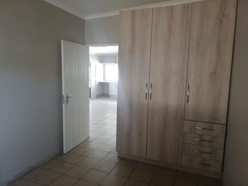 A clean, empty bedroom featuring light wood-finish built-in cupboards, neutral wall tones, and tiled flooring, with a view into an adjacent room.