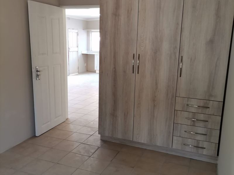 A clean, empty bedroom featuring light wood-finish built-in wardrobes, tiled flooring, and neutral wall tones, with a view into an adjacent room.