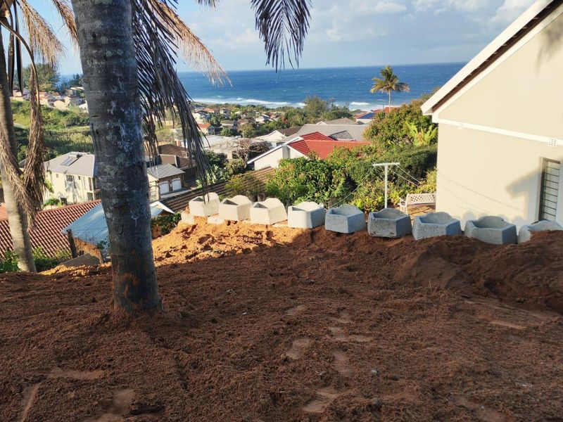 Rear exterior view of the property featuring a sloped, unlandscaped yard with retaining wall blocks and a scenic coastal ocean view.