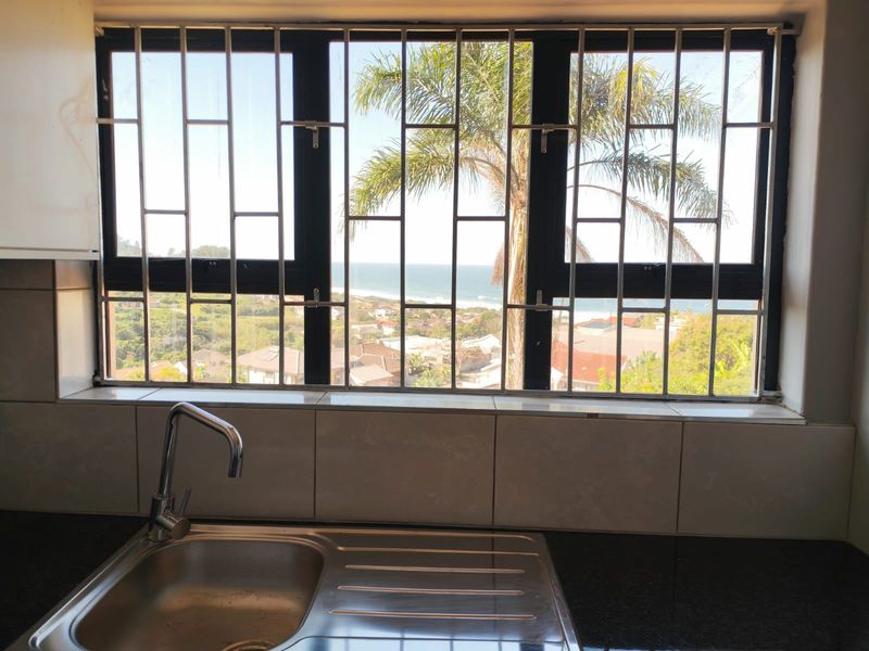 A kitchen sink area featuring a stainless steel basin, mixer tap, and tiled splashback, with a window offering scenic coastal views.