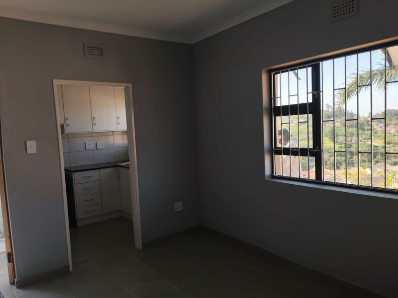 An empty open-plan living area featuring tiled floors and a view through security-barred windows, with an adjacent kitchen area visible through the doorway.