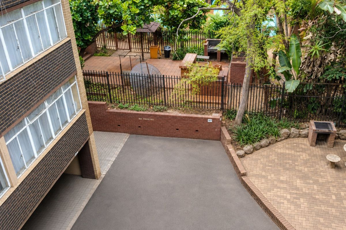 Elevated view of the apartment complex rear exterior, featuring a paved courtyard, built-in brick braai area, and secure perimeter fencing.