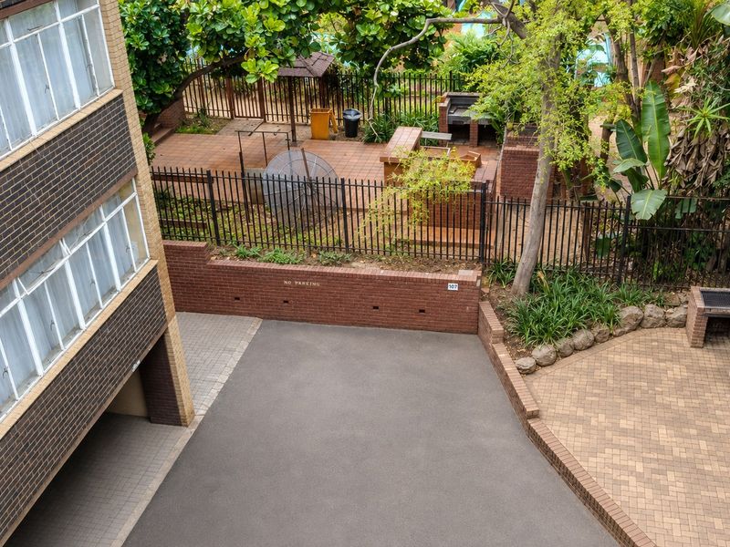 Elevated view of the apartment complex rear exterior, featuring a paved courtyard, built-in brick braai area, and secure perimeter fencing.