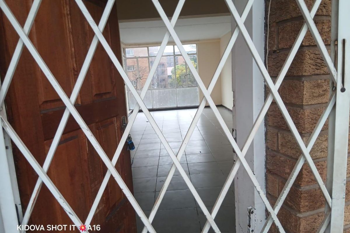 An empty, open-plan living area featuring tiled flooring and large windows, viewed through a security gate.