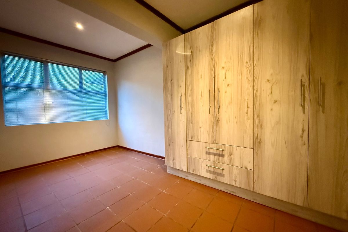 A clean, empty bedroom featuring light wood-finish built-in cupboards, terracotta-style tiled flooring, and a window with fitted blinds.