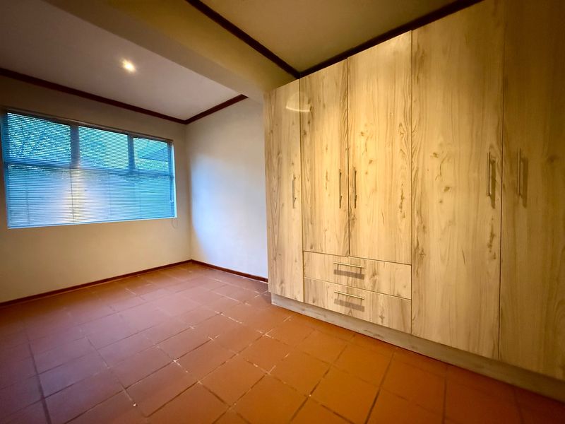 A clean, empty bedroom featuring light wood-finish built-in cupboards, terracotta-style tiled flooring, and a window with fitted blinds.