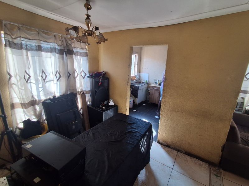 Open plan living area leading into a small kitchen space, featuring tiled floors and dated wall finishes requiring maintenance and cosmetic updates.