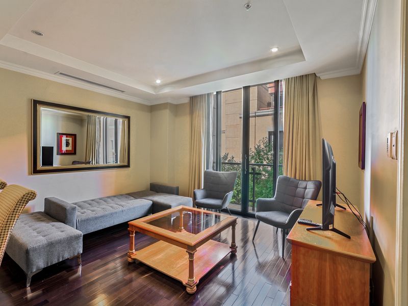 A well-lit living room featuring dark hardwood flooring, a recessed ceiling with cove lighting, and a sliding glass door leading to a balcony.