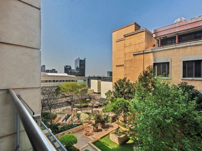 An elevated urban view overlooking a landscaped courtyard and surrounding city buildings from a balcony with a glass balustrade.
