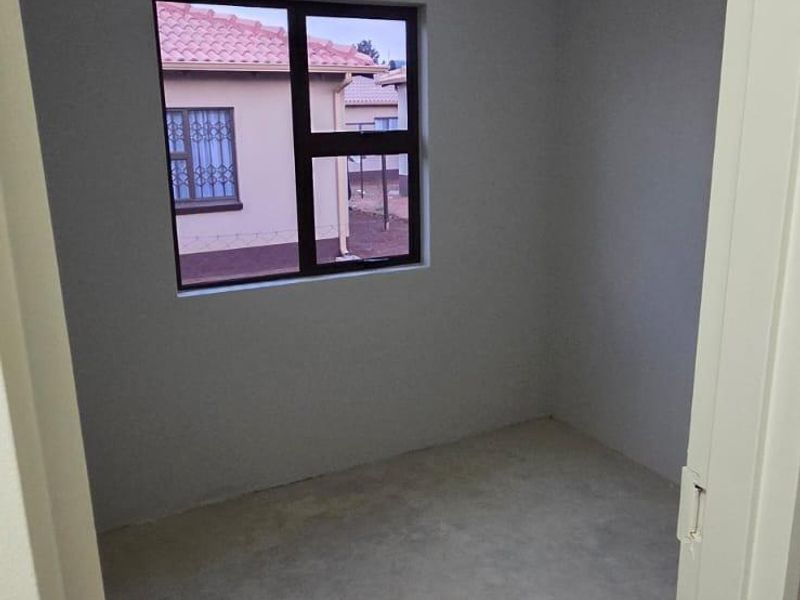 A small, empty bedroom featuring a ceiling light fixture, a window with security bars, and an unfinished concrete floor.