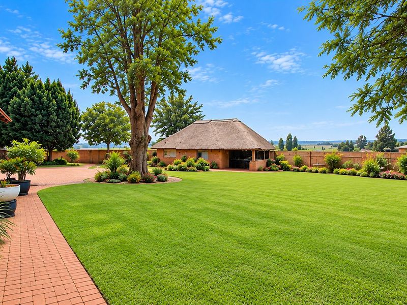 A well-maintained expansive garden featuring a manicured lawn, brick pathways, and a traditional thatched-roof outdoor entertainment area.