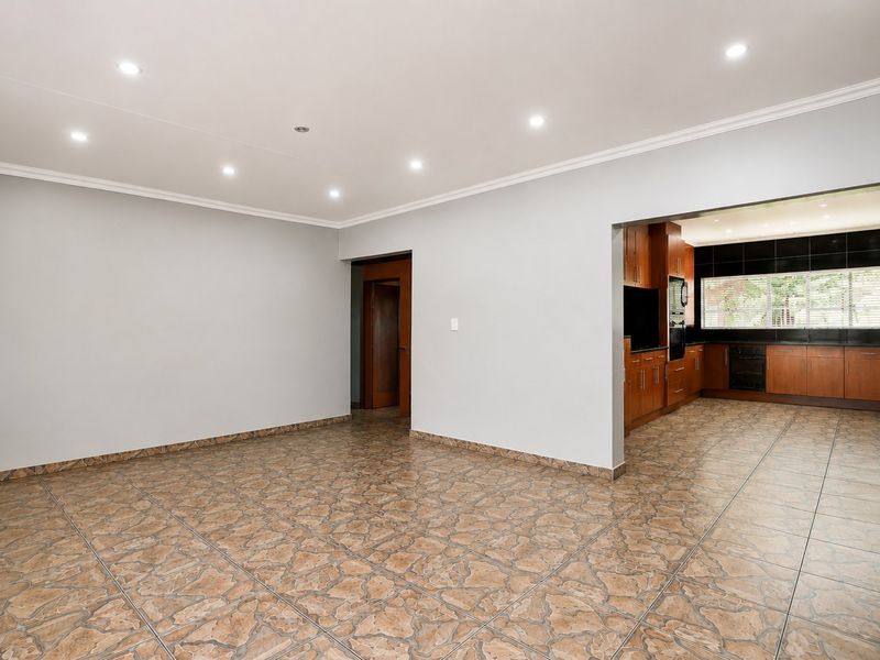 An open-plan living area featuring tiled flooring and recessed lighting, with a view into a kitchen equipped with built-in wooden cabinetry and appliances.