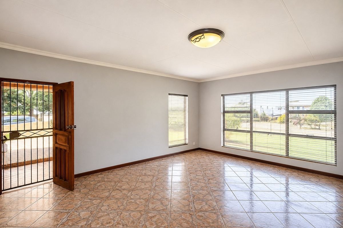 A bright, empty living area featuring tiled flooring, large windows with venetian blinds, and a secure exterior door access.