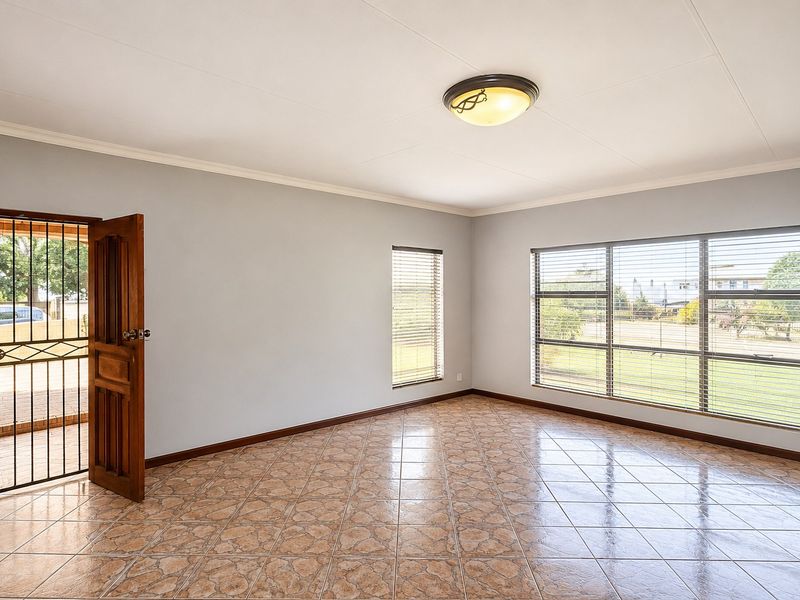 A bright, empty living area featuring tiled flooring, large windows with venetian blinds, and a secure exterior door access.