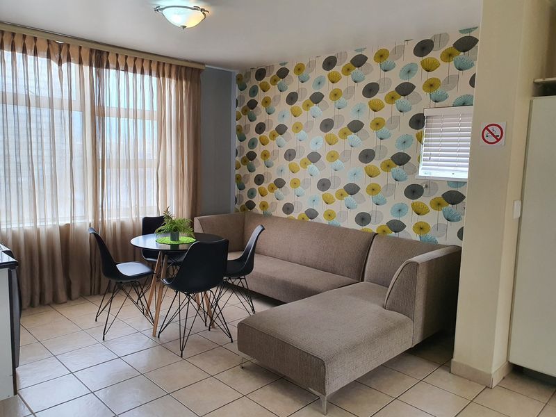 An open-plan living and dining area featuring tiled flooring, patterned feature wallpaper, and large windows with curtain tracks and venetian blinds.