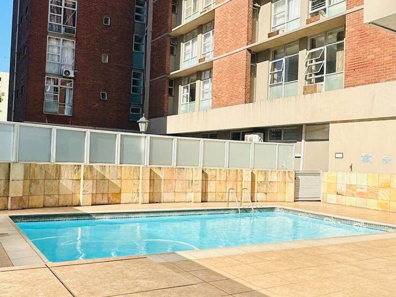 A well-maintained outdoor swimming pool area featuring a tiled deck, stone wall cladding, and privacy screening, set against a multi-story apartment building.