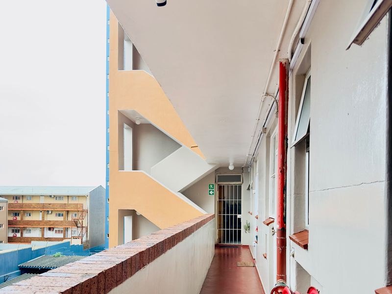 An exterior apartment building walkway featuring a brick parapet, security cameras, and a fire hose reel, providing access to residential units.