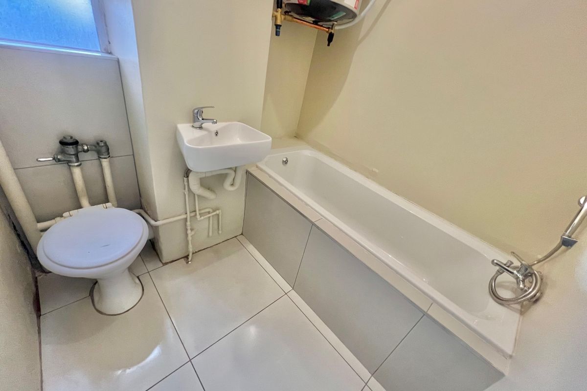 Compact bathroom featuring a bathtub, wall-mounted basin, and toilet, with exposed plumbing and an overhead electric water heater.