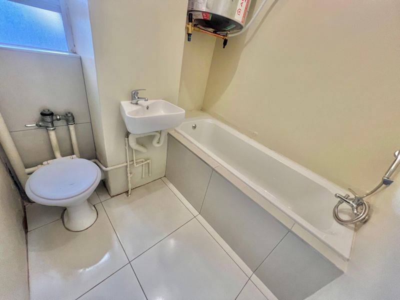 Compact bathroom featuring a bathtub, wall-mounted basin, and toilet, with exposed plumbing and an overhead electric water heater.