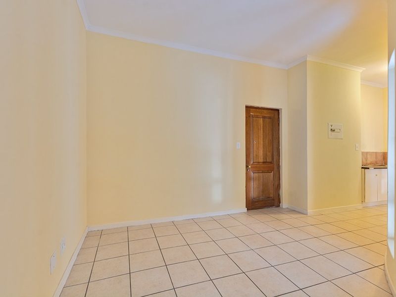 An empty open-plan living area featuring neutral walls, tiled flooring, and a classic wooden door, with a glimpse of the kitchen area beyond.