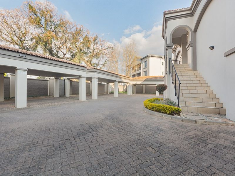 A well-maintained apartment complex exterior featuring covered parking bays, a paved driveway, and a stone staircase leading to the entrance.