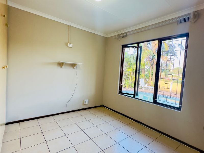 An empty bedroom featuring tiled flooring, a built-in cupboard, and a window with security bars overlooking the pool area.