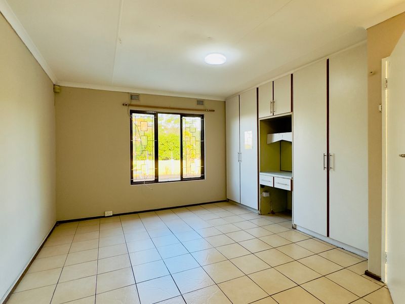 A spacious, empty bedroom featuring neutral tiled flooring, built-in cupboards with integrated vanity space, and a window with security bars.