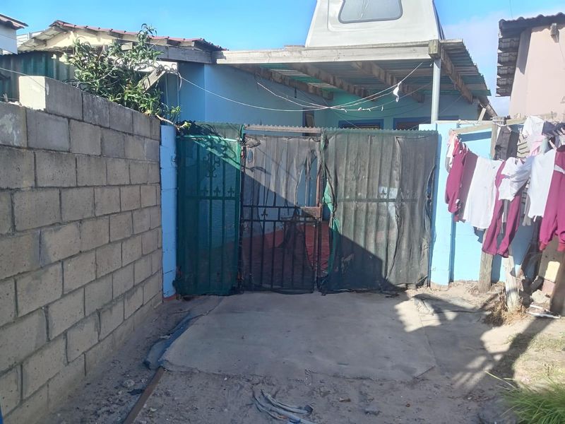 Rear exterior view showing a concrete block boundary wall, metal security gate, and a basic covered patio area with weathered finishes.