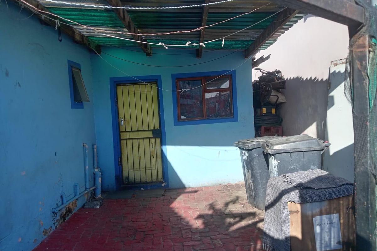 A rear exterior area featuring a paved courtyard, corrugated roof covering, and a security door, showing signs of significant wear and maintenance requirements.