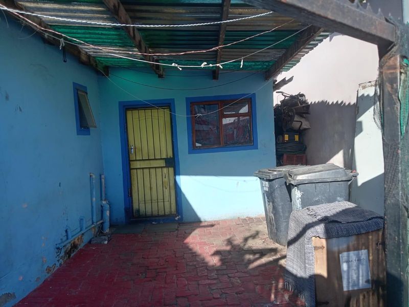 A rear exterior area featuring a paved courtyard, corrugated roof covering, and a security door, showing signs of significant wear and maintenance requirements.