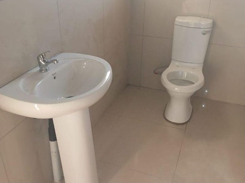 A clean, minimalist bathroom featuring a pedestal washbasin and a standard toilet, finished with neutral ceramic wall and floor tiles.
