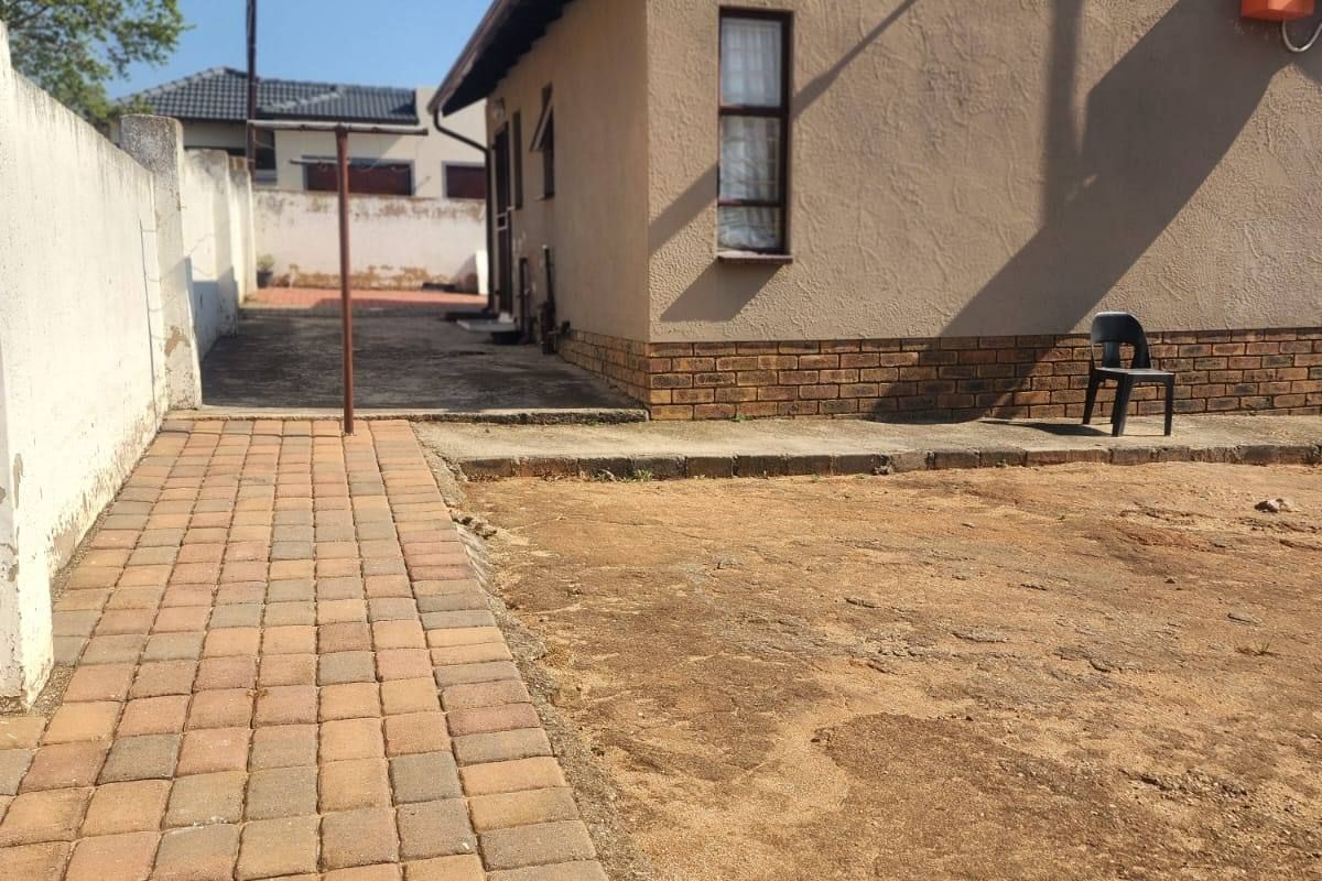 Rear view of the property featuring a paved walkway, plastered exterior walls with brick plinth, and an open, unlandscaped garden area.