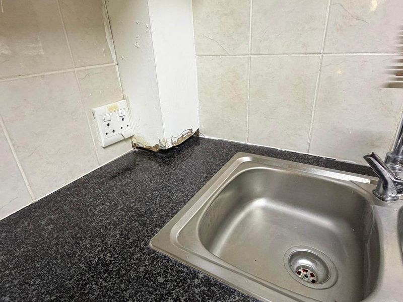 A close-up view of a kitchen sink area featuring a granite countertop, stainless steel basin, and tiled backsplash with visible wear at the wall junction.