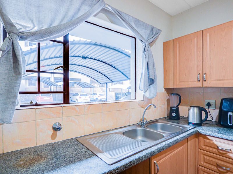 A functional kitchen area featuring built-in wooden cabinetry, a double bowl stainless steel sink, and tiled backsplash with a view of the parking area.