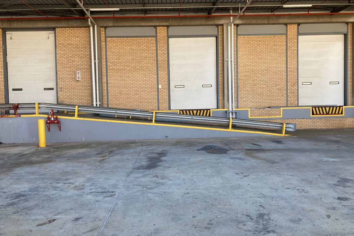 Industrial loading bay area featuring multiple roller shutter doors, a concrete ramp with safety railings, and a durable brick facade.