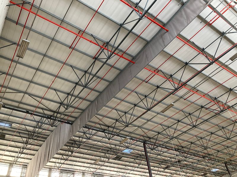 View of an industrial warehouse ceiling featuring exposed steel roof trusses, integrated fire sprinkler piping, and industrial lighting fixtures.
