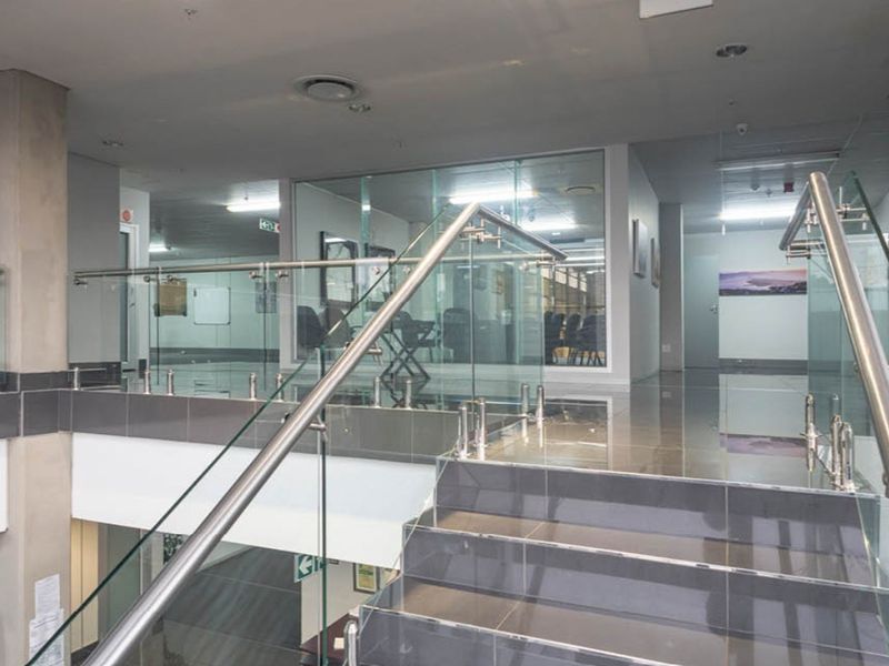 Modern industrial office hallway featuring a glass balustrade staircase, polished tile flooring, and glass partition walls creating an open and professional workspace environment.