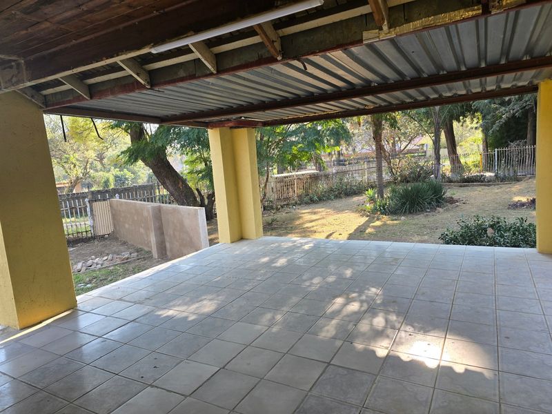 A covered outdoor patio area featuring tiled flooring, exposed timber rafters, and concrete support pillars, overlooking a spacious garden.